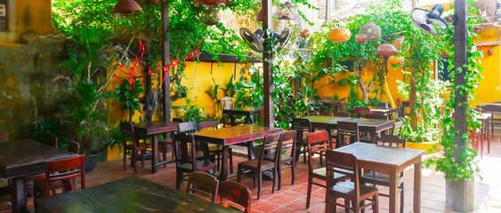 Panorama view lush vines, flowering shrubs along alleys and courtyards garden-style restaurant creating cozy, semi-private nooks for dining in Hoi An, rustic wooden tables and ceiling fans