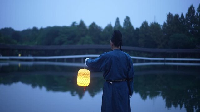 Man in traditional robe holding a glowing lantern by the lakeside at dusk, back view with serene reflection on water