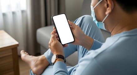 Man wearing a face mask sitting on a couch using a smartphone with a blank white screen to display content