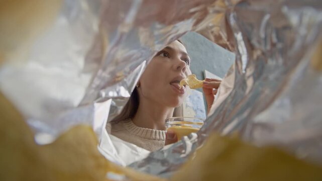 POV bottom view shot of young Caucasian woman enjoying crispy nachos while watching film at home, taking chips from package and dipping in creamy cheese sauce