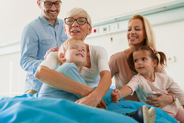 Obraz premium Smiling Caucasian Family Visiting Grandmother in Hospital Room Sharing Warm Moments with Kids and Adults