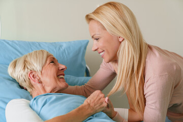 Blonde Woman Visiting Elderly Mother in Hospital Smiling Warmly in a Bright Indoor Room