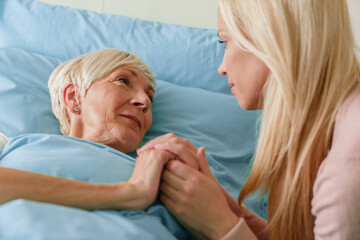 Fototapeta premium Blonde Woman Comforting Elderly Grandmother in a Hospital Room with Warm Lighting and Emotional Connection