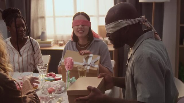 Blindfolded young spouses guessing objects without seeing them during contest at festive table held by their friends at pink and blue party, they removing blindfolds and high-fiving each other