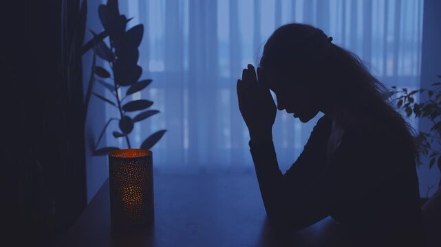 Portrait adult woman praying whispers words of gratitude, hands folded in prayer. Dark silhouette female prayer. Girl Christian praying to God concept of religion faith hope blessing. home room window
