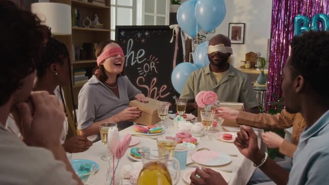 Blindfolded diverse young couple taking part in contest at holiday table with multiethnic friends watching, they taking items for future baby out of boxes and trying to guess at gender reveal party