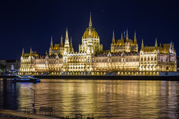 Fototapeta premium Night panorama of Hungarian Parliament from Danube's opposite bank, showcasing neo-Gothic architecture with towers, spires, and dome illuminated against indigo-black sky. Budapest, Hungary.
