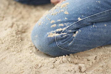 A person kneels in soft sand, showcasing jeans marked with grains. The warm sunlight highlights the relaxed atmosphere, hinting at a day of leisure