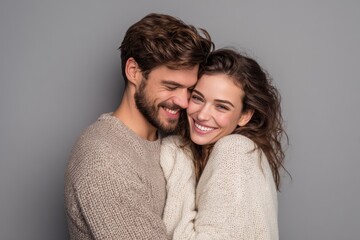 Couple sharing a joyful moment while embracing in cozy sweaters, showcasing tenderness and happiness in a soft gray backdrop during a casual indoor setting