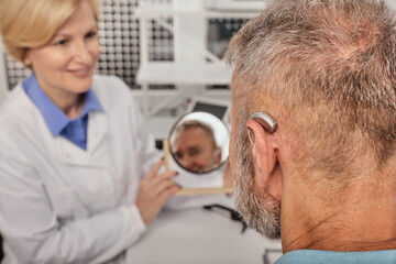 Senior man with BTE hearing aid looks at himself in mirror held by smiling audiologist, and tries...