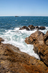 Waves crashing on rugged coastal rocks with turquoise sea and clear blue sky on sunny day