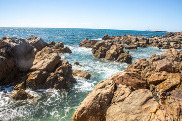 Waves crashing against rugged coastal rocks under clear blue sky on a bright sunny day, natural seaside landscape