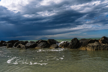 Rocks line the shore as a storm gathers on the horizon, waves crashing softly. Nature's display of power and serenity.