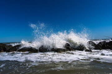 Obraz premium Waves crashing against a rocky shoreline under a vibrant blue sky, creating a dynamic coastal scene with powerful sea spray.