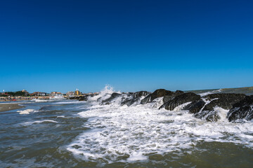 Obraz premium Waves crashing against rocks near a beach, creating a dynamic seascape against a clear blue sky. Coastal beauty unfolds.