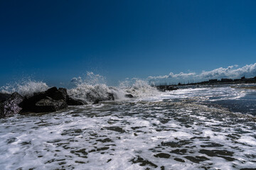 Waves crash against rocks on a sunny day, creating sea foam. The sky is blue, and the horizon shows city buildings.