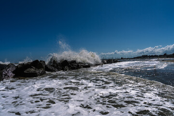 Ocean waves crash against a rocky shoreline, creating a dynamic scene under a clear blue sky near the city.