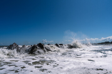 Waves crash against rocks under a clear blue sky, creating a dynamic scene of coastal energy. The shoreline meets the open water.