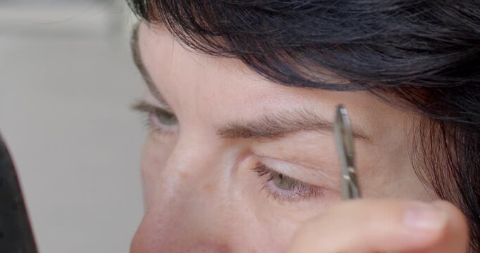 A woman meticulously performs eyebrow grooming with tweezers, emphasizing the importance of precision and skill in maintaining perfectly shaped eyebrows for an elegant look