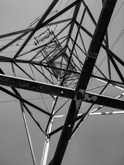Monochrome worm's-eye view of a tall electrical pylon, highlighting the intricate, abstract geometric pattern of its metal structure against the sky