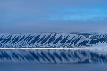 Snow covered mountains of Svalbard reflected in the still Arctic fjord, capturing the serene and...