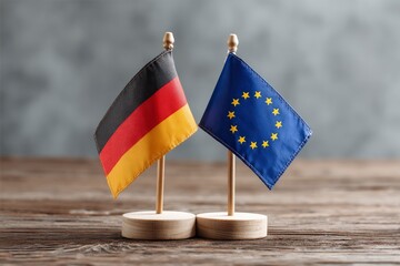 Two flags Germanys black red and gold and the EUs blue with yellow stars stand on a wooden table against a blurred gray background