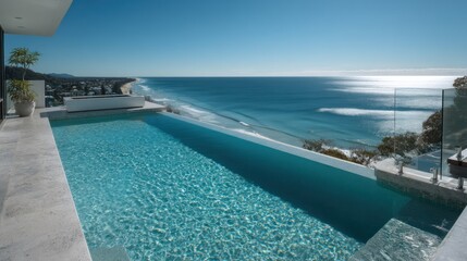 Fototapeta premium A stunning infinity pool extends dramatically over a cliffside, overlooking a brilliant blue ocean. The pool?s turquoise water reflects the sky and sunlight, creating a shimmering effect. The pool?