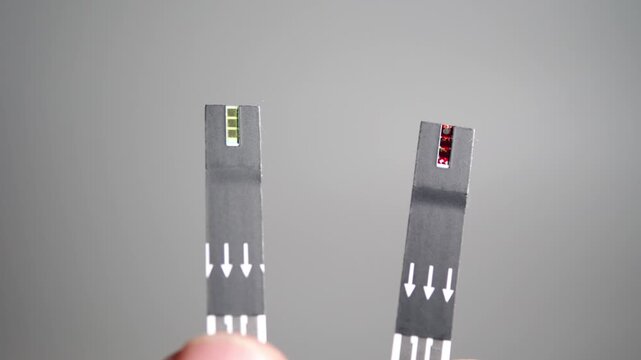 A hand holds two blood glucose test strips, one with a drop of blood on its tip