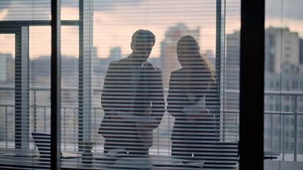 Two confident partners discussing contract papers standing at office window