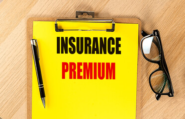 Words insurance premium written on yellow clipboard with pen and eyeglasses on wooden desk