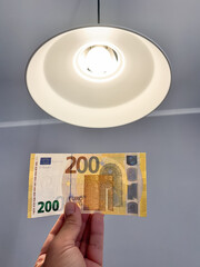 Hand holding a 200 euro banknote under a modern ceiling light, symbolizing inflation and...