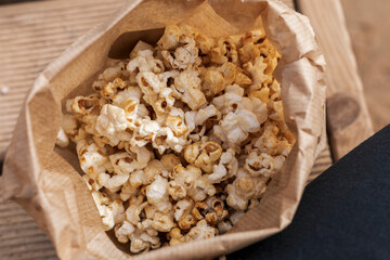 A brown paper bag filled with freshly popped popcorn is resting on a wooden surface. The crunchy snack is perfect for a cozy movie night, inviting everyone to indulge and enjoy
