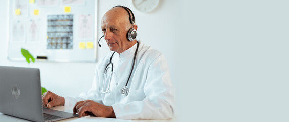 Elderly senior male doctor in white coat with stethoscope using laptop talking video conference call with patient at hospital or clinic interior. Consulting and therapy