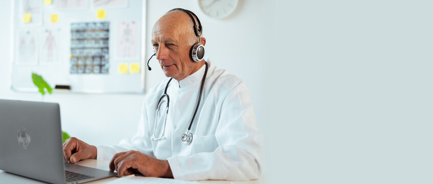 Elderly senior male doctor in white coat with stethoscope using laptop talking video conference call with patient at hospital or clinic interior. Consulting and therapy