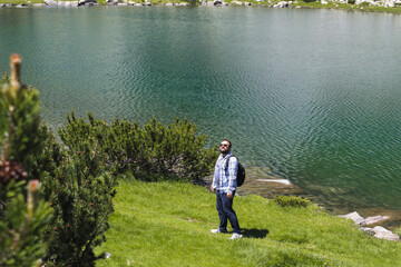 Bearded hiker by a clear turquoise mountain lake and grassy shore, under a sunny sky in tranquil...