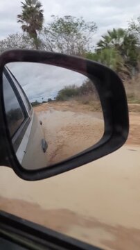 pov: retrovisor de carro em estrada sem pavimenta&ccedil;&atilde;o com po&ccedil;as de barro  