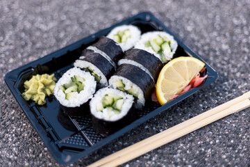 A tray holds several fresh cucumber sushi rolls, paired with a dollop of wasabi and a slice of lemon. The setting features a textured surface, emphasizing the vibrant colors of the dish