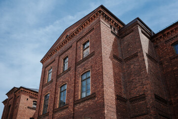 A low-angle photograph of a massive, historic industrial building made from dark red brick, set against a blue sky with clouds.