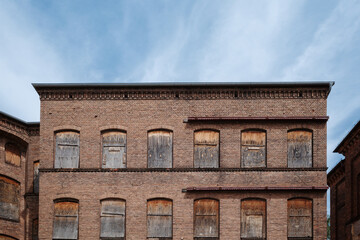 The weathered brick facade of an old, abandoned industrial factory or mill under a blue sky. All of the arched windows on the upper floors have been boarded up with old wood