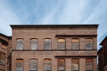 Fototapeten Gebäude In Trümmer The weathered brick facade of an old, abandoned industrial factory or mill under a blue sky. All of the arched windows on the upper floors have been boarded up with old wood  © Peeradontax