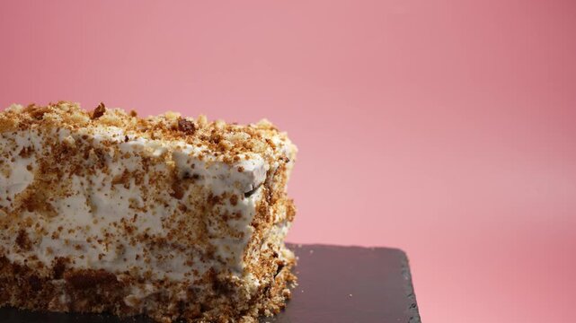A cake slice with white cream and crumbs rotates on a slate plate against a pink background. The video captures its detail and tempting texture.