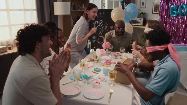 Diverse friends supporting two blindfolded young people trying to guess objects, pregnant woman filming video of them, company raising glasses during pink and blue party in apartment
