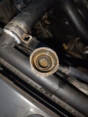 Close-up view of a rusty radiator cap, connected to hoses, in an engine compartment.