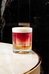 Colorful layered cocktail with foam served in a crystal glass, garnished with a dried citrus slice. Elegant drink presentation on a marble table in a stylish modern bar