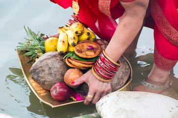 A woman performing Chhath Puja ritual by holding a dala (basket) filled with fruits, coconut, and...