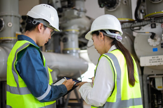 Two engineers in safety gear discussing plans in an industrial facility with machinery in the background