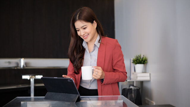 Smiling Asian businesswoman holding a coffee cup while using a digital tablet at a modern glass desk.