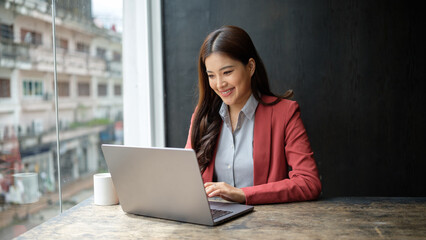 Professional businesswoman in a red blazer working on a laptop by a window with an urban city view.