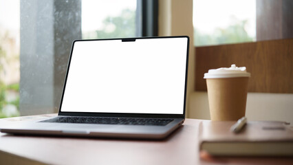 Fototapeta premium Laptop with blank white screen on wooden desk beside coffee cup and notebook in a cozy cafe.
