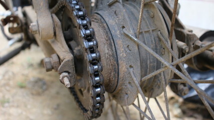 Closeup of a motorcycle rear sprocket and chain system, showing worn metal parts, rusted spokes,...